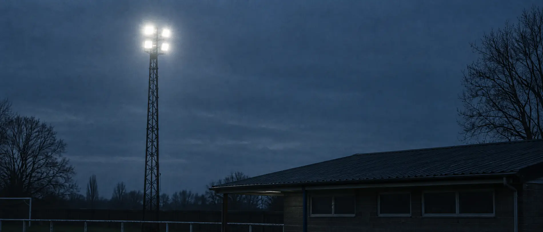 Ein Flutlichtmast und das flache Dach eines Vereinsheims vor tiefblauem Abendhimmel, die Lampen gerade eingeschaltet.