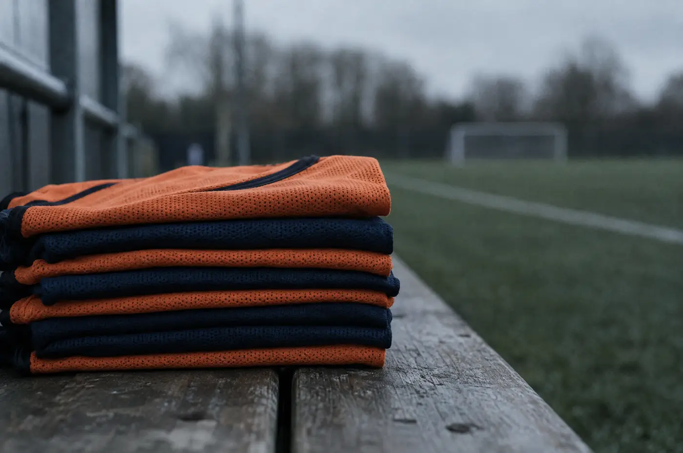 Ein sauber gefalteter Stapel oranger und dunkelblauer Trainingsleibchen auf einer Holzbank am Platzrand.
