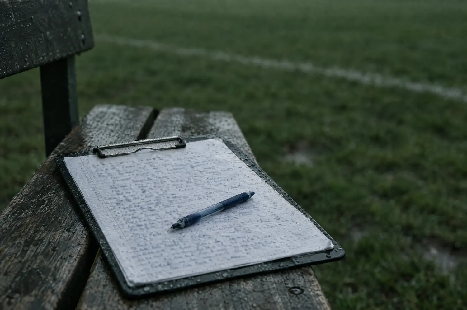 Ein regennasses Klemmbrett mit handschriftlicher Liste und Kugelschreiber auf einer Holzbank am Spielfeldrand.